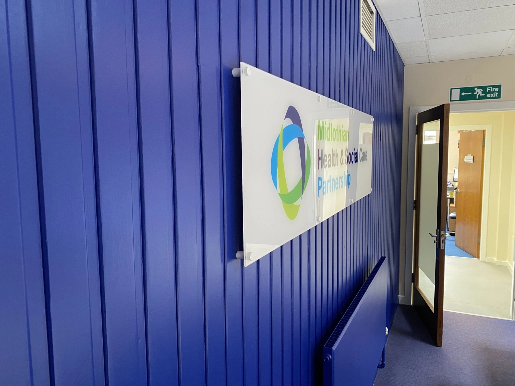 Acrylic stand-off sign on metal fixings mounted on a blue clad wall beside an entrance
