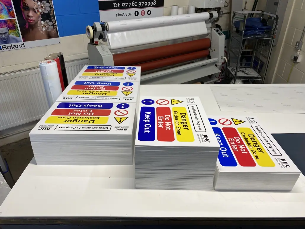Stacks of printed site safety signs on the workshop bench beside the laminator
